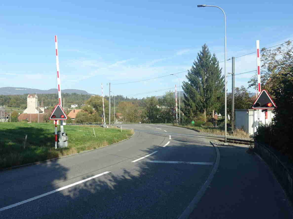 Bahnübergang Jurastrasse mit geöffneten rot-weissen Schranken beidseits der Fahrbahn und dreieckigen Blinklichtsignalen mit rotem Licht. Die asphaltierte Strasse mit Mittellinie führt in einer Kurve über die Gleise, welche eine Fahrstrasse ausserorts erkennen lassen. Im Hintergrund ein Turm, Häuser und bewaldete Hügel, keine Personen oder Fahrzeuge sichtbar.