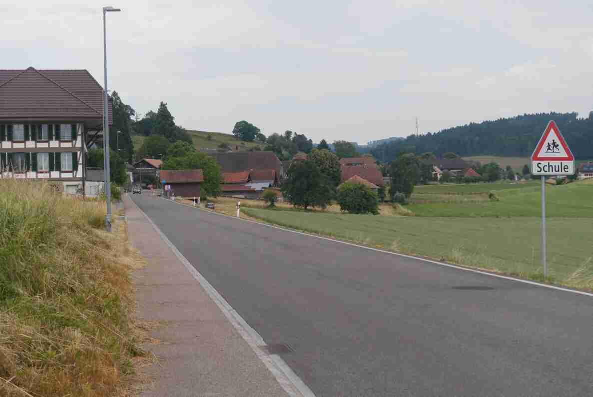 Dorfstrasse in Alchenstorf mit Blick auf ein Bauernhaus mit Riegelfassade links und weitere Liegenschaften mit roten Ziegeldächern im Hintergrund, umgeben von Wiesen und bewaldeten Hügeln. Rechts steht ein Warnschild «Schule» mit Kindersymbolen. Die zweispurige, asphaltierte Strasse ist frei von Verkehr, ein einzelnes Fahrzeug nähert sich in der Ferne. Ein Trottoir verläuft entlang der linken Strassenseite.