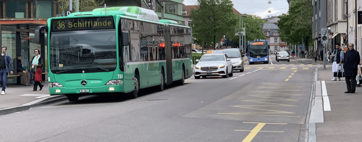 Haltestelle Kinderspital mit grünem Gelenkbus der Linie 36 Richtung Schifflände, der von der Haltebucht in den Verkehr einbiegt. Dahinter folgen Personenwagen, ein Taxi und ein blauer Bus auf der mehrspurigen Strasse mit Tempo-30-Zone-Signal. Fussgänger überqueren die Fahrbahn und warten am Trottoir, Velofahrer sind am Strassenrand sichtbar. Der Asphalt ist trocken und in gutem Zustand.