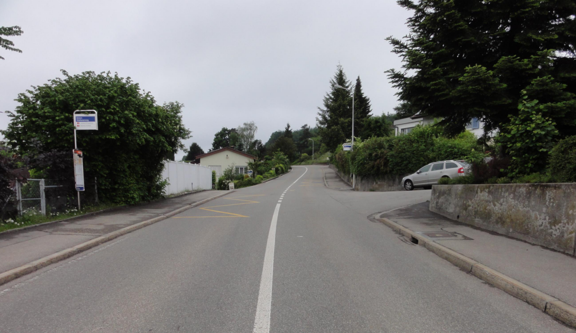 Strassenkreuzung Wallierenweg/Rüttistrasse in Bellach mit Bushaltestelle Wallierenweg links am Strassenrand, eingerahmt von hohen Hecken und Bäumen, die die Sicht auf die einmündende Rüttistrasse einschränken. Der Asphalt weist gelbe Warnmarkierungen und eine Mittellinie auf. Ein parkiertes Auto steht rechts am Strassenrand vor einer Stützmauer. Kein Verkehrssignal oder Messgerät sichtbar, keine Personen im Bild.