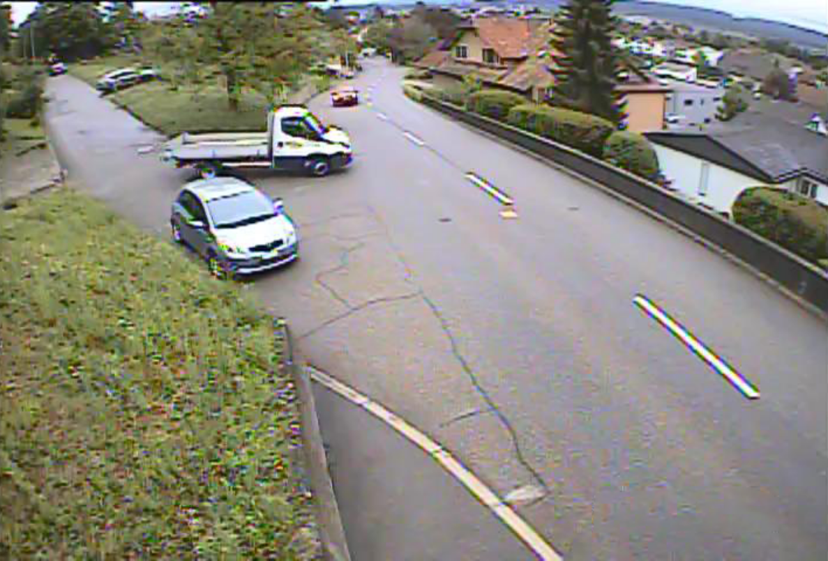 Strassenkreuzung an der Lommiswilerstrasse in Bellach mit Blick auf eine leichte Kurve. Ein Lastwagen mit offener Ladefläche biegt von einer Seitenstrasse ein, ein silberner Personenwagen ist ebenfalls im Einmündungsbereich unterwegs, ein weiteres Auto fährt weiter oben auf der Hauptstrasse. Die Fahrbahn ist zweispurig mit unterbrochener Mittellinie, seitlich begrenzt durch Grünstreifen, Hecken und Wohnhäuser.