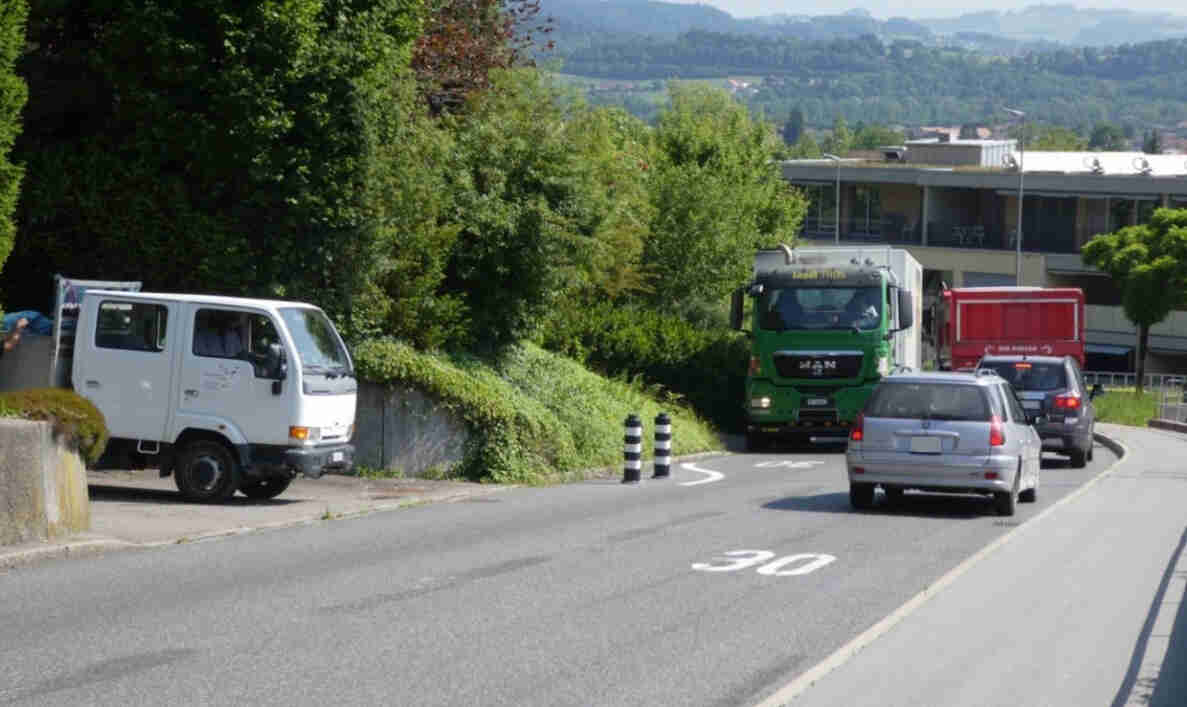 Strasse mit aufgemalter Tempo-30-Markierung, verengt durch schwarz-weisse Leitpoller. Ein grüner Lastwagen und ein weisser Lieferwagen begegnen sich in der Engstelle, dahinter folgen ein silberner Personenwagen, ein weiterer Lastwagen und ein SUV. Die Fahrbahn ist asphaltiert und in gutem Zustand, im Hintergrund ein Gebäude sowie hügelige Landschaft.
