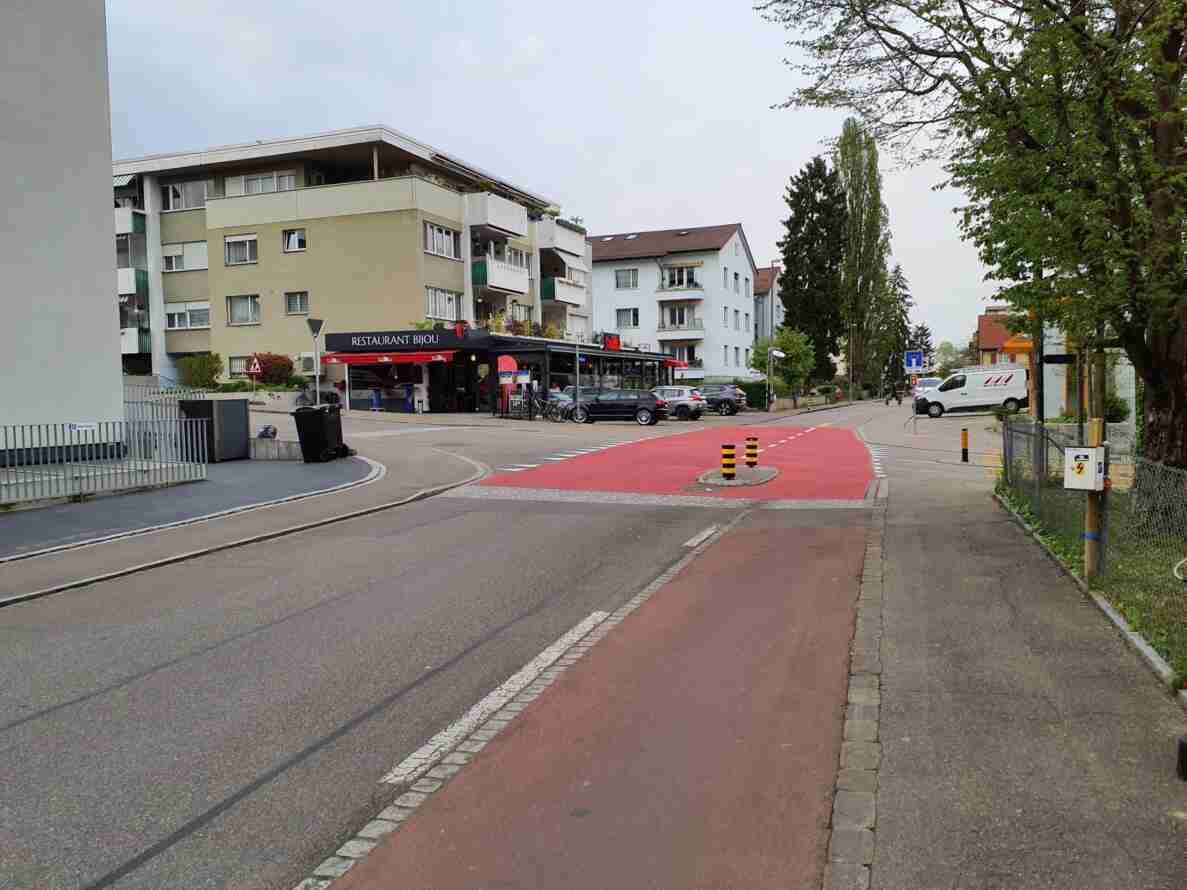 Strassenkreuzung mit rot markiertem Radweg und Fussgängerstreifen vor dem Restaurant Bijou. Verkehrsinsel mit gelb-schwarzen Leitpfosten trennt Fahrbahn und Radweg. Rechts Einbahnstrassen-Signal, links Vortrittssignal. Wohnhäuser im Hintergrund, geparkte Autos und ein Lieferwagen am Strassenrand, Velofahrer auf der Fahrbahn. Asphalt in gutem Zustand mit klaren Fahrbahnmarkierungen.