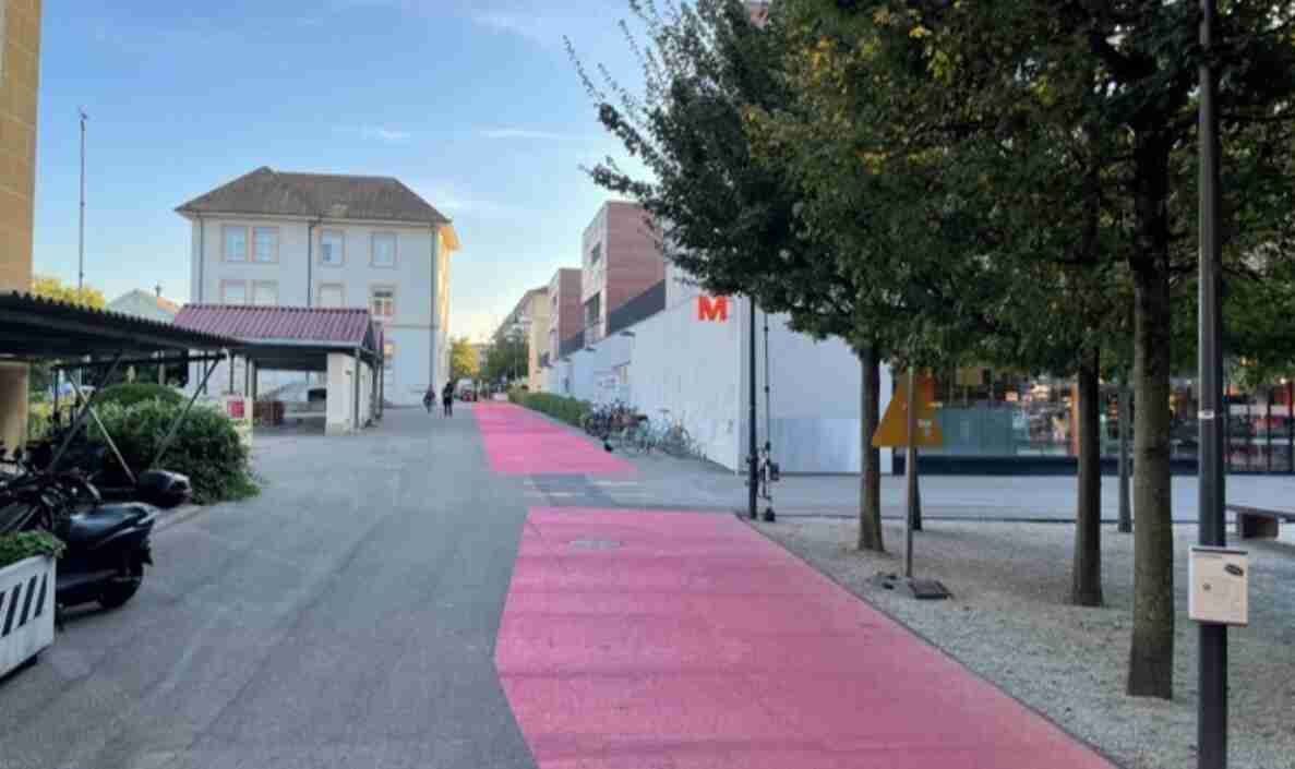 Dorfplatz Binningen mit pink markierter kantonaler Veloroute, die geradeaus durch den Platz führt. Rechts stehen Bäume und ein Fahrradständer mit parkierten Velos, links ein überdachter Unterstand mit abgestellten Motorrollern. Im Hintergrund gehen Personen zu Fuss auf dem Platz. Die breite, verkehrsberuhigte Fläche dient gemeinsam Fussgängern und Velofahrenden, umgeben von Wohn- und Geschäftshäusern.