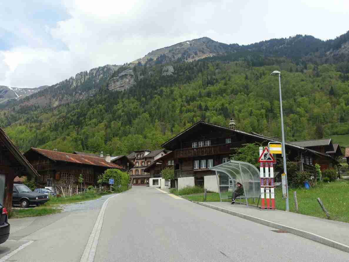 Brünigstrasse in Brienzwiler mit intaktem Belag und Mittellinie, gesäumt von traditionellen Schweizer Holzhäusern vor bewaldeten Berghängen. Rechts eine Postauto-Haltestelle mit Wartehäuschen, auf dessen Bank eine sitzende Person wartet. Daneben ein Baustellen-Warnsignal mit rot-weissen Streifen. Die Signalisation und Bushaltestelle deuten auf eine überörtliche Verbindungsstrasse hin. Am Strassenrand parkierte Fahrzeuge.