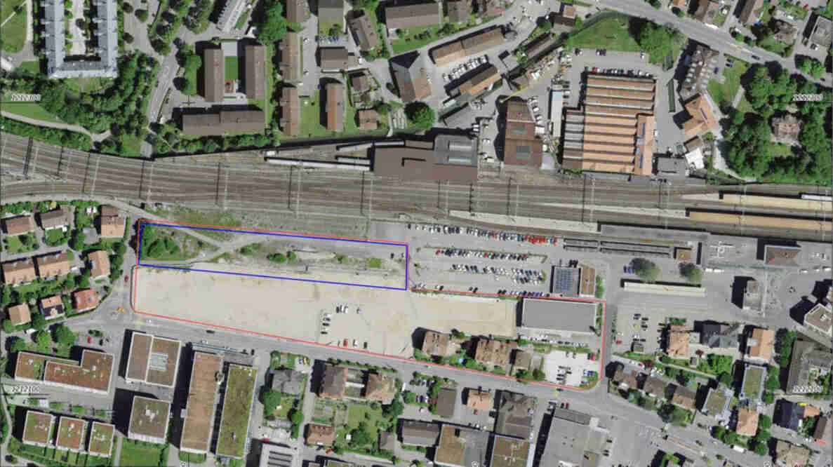 Luftbild des Aebiareals in Burgdorf mit markierten Parzellengrenzen entlang der Bahngleise. Sichtbar sind ein grosses Bahnhofsareal mit mehreren Gleisen, angrenzende Gewerbe- und Wohngebäude, Parkplätze mit Fahrzeugen sowie ein unbebautes, teils asphaltiertes Areal. Umliegende Strassen erschliessen die Quartiere für den Fahrzeug- und Fussverkehr.
