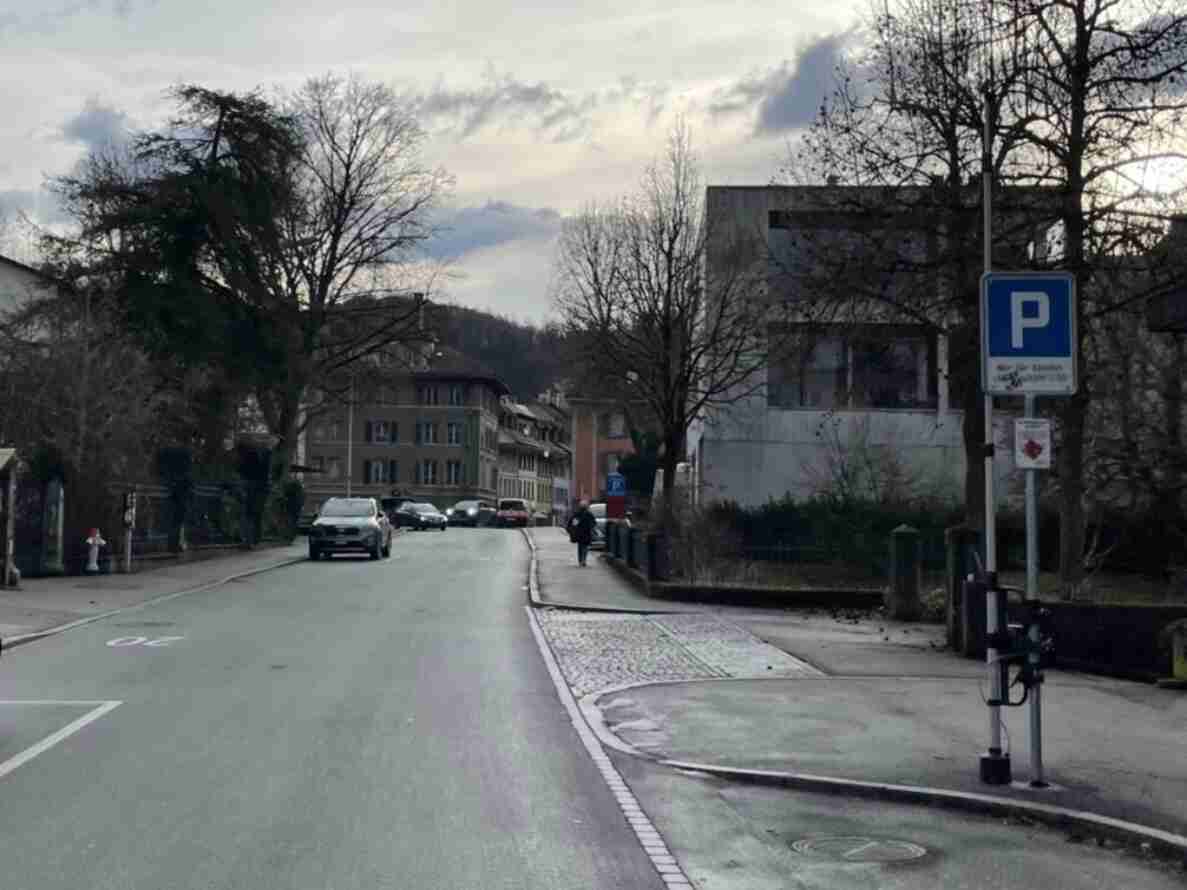 Blick entlang der Lyssachstrasse mit trockener Fahrbahn, aufgemalter Geschwindigkeitsbeschränkung 20 auf dem Asphalt sowie einem Pflastersteinstreifen als Querungshilfe. Am Strassenrand parkierte Fahrzeuge, ein Parkschild und ein gehender Mensch auf dem Trottoir. Kahle Bäume säumen die Strasse, im Hintergrund mehrstöckige Wohnhäuser und ein bewaldeter Hügel.
