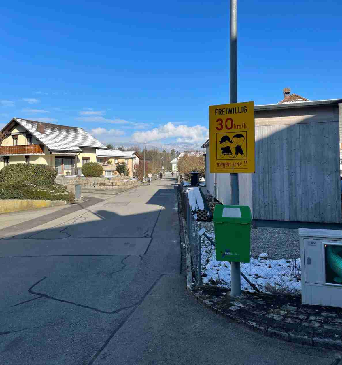 Gemeindestrasse in Dotzigen mit gelbem Hinweisschild «Freiwillig 30 km/h wegen uns» mit Kinderpiktogramm, das auf eine Tempo-30-Zone hinweist. Die Fahrbahn ist trocken und schneefrei, am Rand liegt noch etwas Schnee. Im Hintergrund fahren mehrere Velofahrende auf der Strasse, ein Briefkasten und ein grüner Robidog-Kübel stehen am Wegrand.