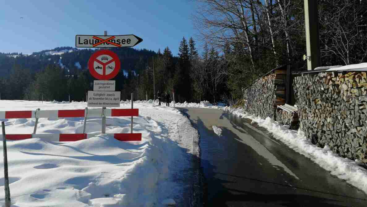 Wegweiser Richtung Lauenensee mit durchgestrichenem Ortsnamen, darunter Verbotssignal für Motorwagen, Motorräder und Motorfahrräder mit Zusatztafel «Zubringerdienst gestattet» sowie Gültigkeitshinweis von November bis ca. April. Eine rot-weisse Barriere steht am Strassenrand. Die schneegeräumte, teils nasse Strasse ist frei befahrbar, im Hintergrund gehen zwei Personen am Wegrand, rechts stapelt sich Brennholz.