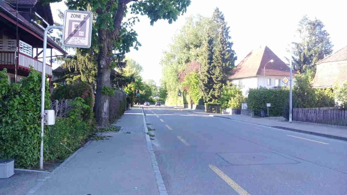 Strasse durch Wohnquartier mit Schild «Ende Zone 2» für Parkierungsbeschränkung, gelber Sicherheitslinie in Strassenmitte und Trottoirs beidseits. In der Ferne zeigen zwei Ampeln Grün für den Verkehr, zwei Personenwagen nähern sich auf der Fahrbahn. Ein Gerät zur Messung ist am Signalmast angebracht. Einfamilienhäuser mit Gärten säumen die Strasse, die einwandfreien Asphaltbelag aufweist.