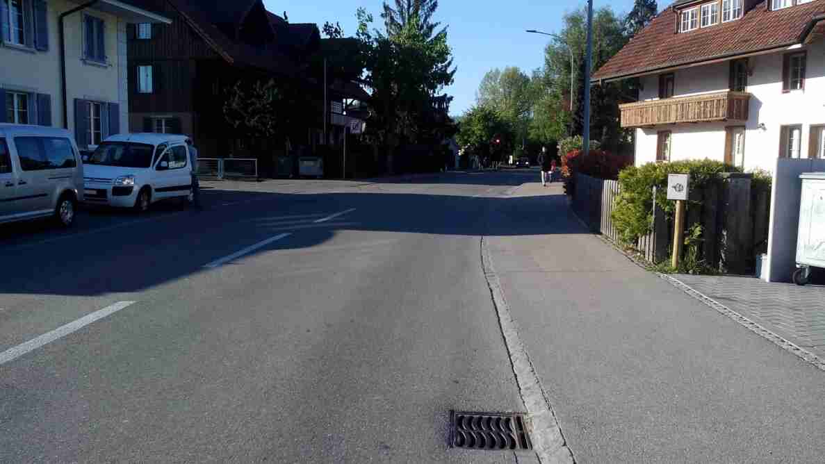 Aarbergstrasse in Lyss, Blick Richtung Süd, mit trockenem Asphaltbelag und Trottoirs beidseitig. Am Strassenrand links parkierte Fahrzeuge, eine Person steht dahinter. Im Hintergrund zeigt eine Ampel Rotlicht, was auf eine Verkehrsregelung hinweist. Rechts gehen zwei Fussgänger dem Trottoir entlang. Umgebende Gebäude im traditionellen Schweizer Baustil säumen die Strasse.