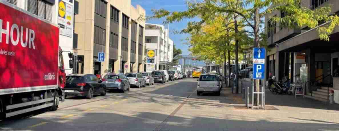 Bahnhofstrasse in Lyss mit beidseitig parkierten Fahrzeugen entlang eines markierten Parkstreifens. In der Strassenmitte wartende Autos vor einer Lichtsignalanlage, ein gelber Bus nähert sich von vorne. Ein blaues Parkschild mit Zusatztafel steht rechts am Trottoirrand, daneben ein parkiertes Motorrad. Links ein Lastwagen mit rotem Werbeaufdruck. Bäume säumen die Strasse, Fussgänger sind keine sichtbar.