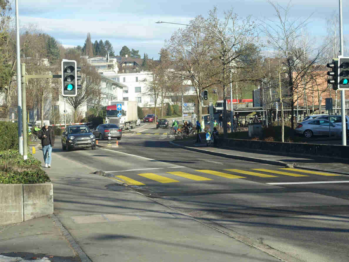 Bürenstrasse in Lyss mit gelb markiertem Fussgängerstreifen, an dem Schulkinder mit Leuchtwesten und Begleitperson auf der Verkehrsinsel warten. Ampeln zeigen beidseitig Grünlicht für den Fahrzeugverkehr, Lastwagen, Personenwagen und Velofahrende befahren die zweispurige Strasse. Eine Person mit Mütze überquert die Fahrbahn ausserhalb des Streifens. Hinweisschilder zeigen Richtung Kath. Kirche und Spitex Seeland. Fahrbahnbelag ist trocken und intakt.