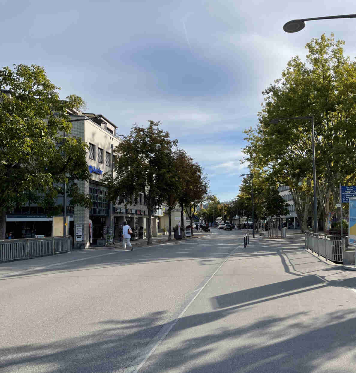 Strasse mit Querungsstelle Lyssbach Brücke im Marktplatzbereich, gesäumt von Bäumen und mehrstöckigen Geschäftshäusern, eines davon mit Schriftzug «Basler». Eine Person überquert zu Fuss die Fahrbahn, im Hintergrund weitere Fussgänger und einzelne parkierte sowie fahrende Autos. Rechts Wegweiser zu Kultur Mühle, Bibliothek und weiteren Zielen. Fahrbahn asphaltiert und trocken, Fussgängerstreifen und Randsteine begrenzen die verkehrsberuhigte Zone im Ortszentrum.
