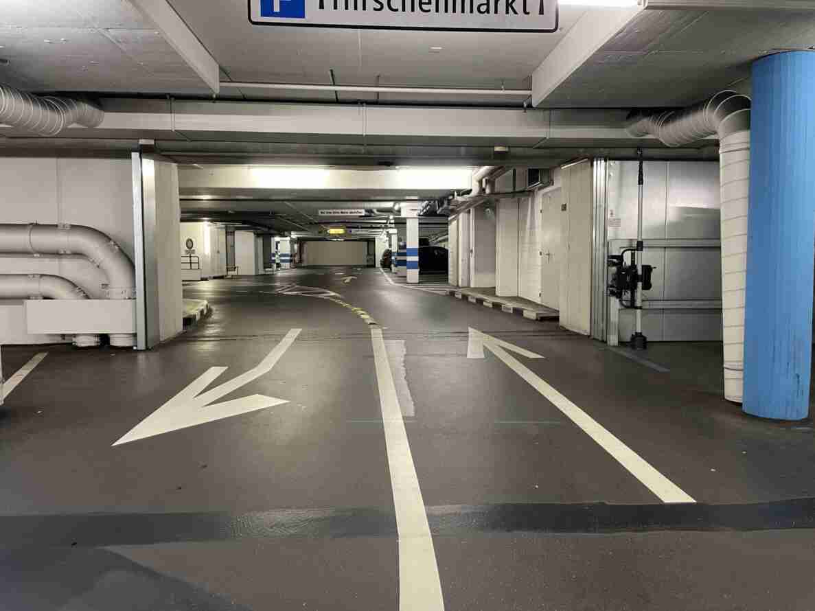 Einstellhalle Hirschenmarkt mit trockener, glatt asphaltierter Zufahrt und aufgemalten Richtungspfeilen für Ein- und Ausfahrt. Ein Hinweisschild «Bei Stau bitte Motor abstellen» sowie gelbe Richtungstafeln hängen von der Decke. An einer Betonsäule rechts ist ein Messgerät zur Verkehrserfassung montiert. Parkfelder mit blau-weisser Markierung sind im Hintergrund sichtbar, keine Personen anwesend.