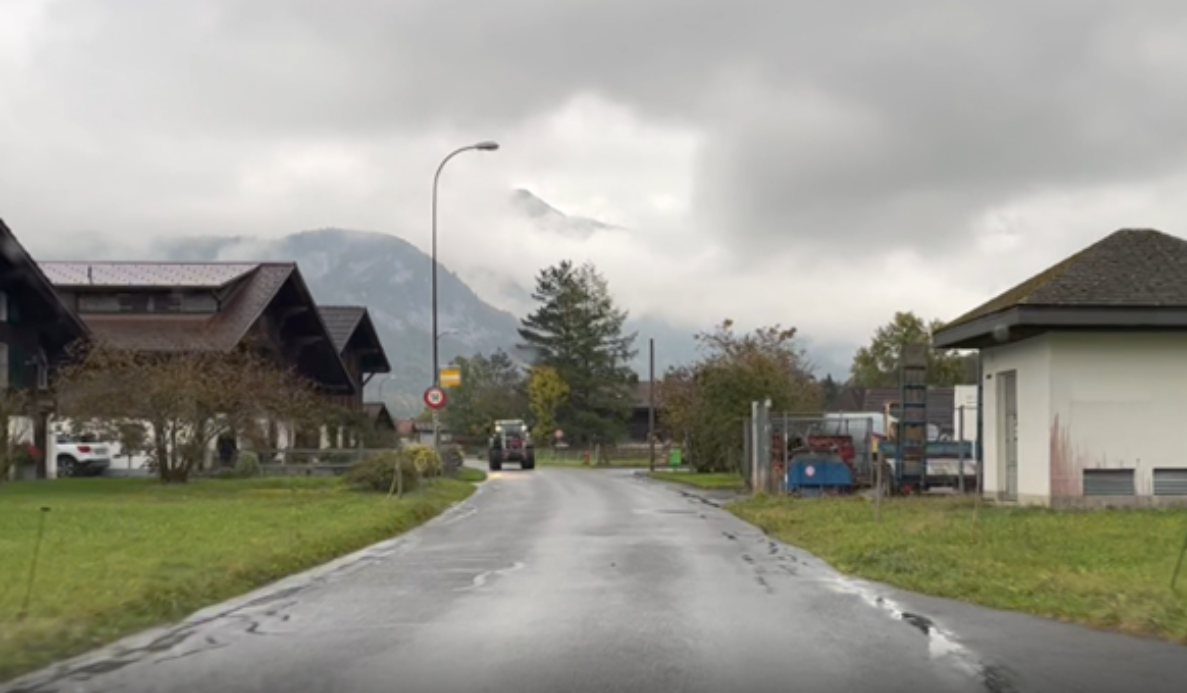Balmstrasse in Meiringen mit schmaler, zweispuriger Fahrbahn zwischen Wohnhäusern im Chalet-Stil und einem Gewerbegebäude. Ein Traktor fährt entgegenkommend auf der Strasse. Am rechten Fahrbahnrand steht ein Verbotssignal für Motorwagen und Motorräder. Grünflächen säumen beidseitig die Strasse, im Hintergrund erheben sich Berge.