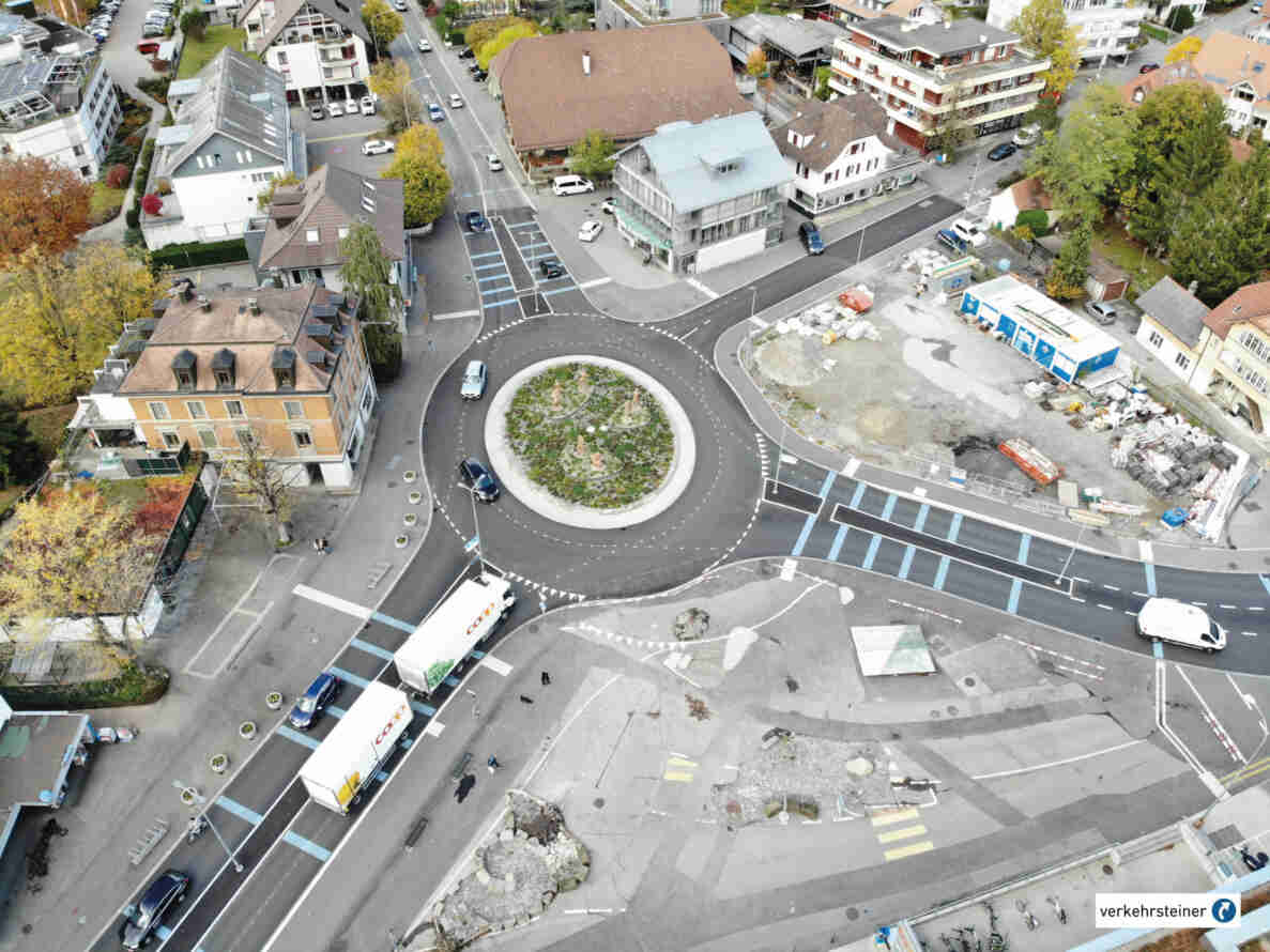Luftaufnahme des neuen Kreisverkehrs in Münsingen mit begrünter Mittelinsel und mehreren zufahrenden Strassen. Der frisch asphaltierte Belag ist mit weissen Leitlinien und blau markierten Parkfeldern versehen, Fussgängerstreifen führen zu den Zufahrten. Mehrere Personenwagen, ein Lieferwagen sowie zwei Coop-Lastwagen befahren die Kreiselarme, während Fussgänger die angrenzenden Gehwege nutzen. Rechts ist eine Baustelle mit Materiallagern sichtbar, eine Tempo-30-Signalisation weist auf eine verkehrsberuhigte Zone hin.