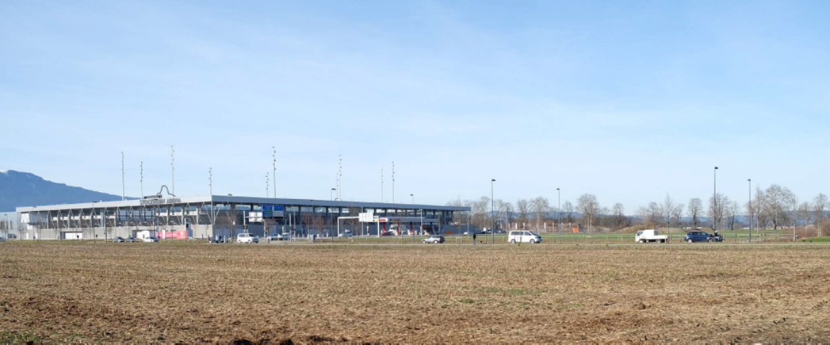 Blick über ein abgeerntetes Feld auf die Zufahrtsstrasse beim Sportcluster Thun Süd, Standort der geplanten neuen Sporthalle. Auf der breiten Strasse mit mehreren Fahrspuren und Trottoir fahren einzelne Personenwagen sowie ein Lieferwagen, eine Person überquert die Fahrbahn. Blaue Wegweisertafeln und Lichtmasten säumen den Strassenraum vor der Sportarena-Halle im Hintergrund.
