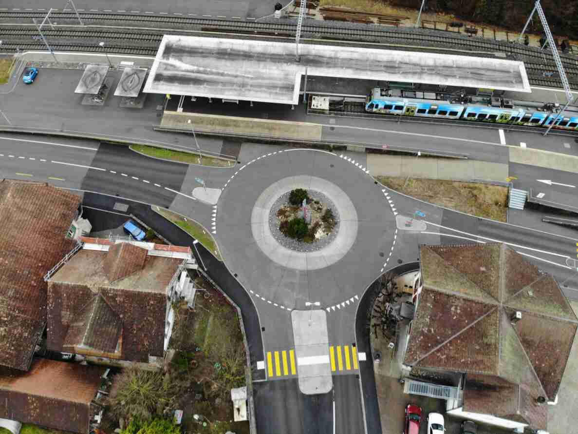 Luftaufnahme des Kreisverkehrs beim Bahnhof Ramsei mit begrünter Mittelinsel, umgeben von Fussgängerstreifen mit Fussgängerschutzinsel. Ein Regionalzug steht am Bahnsteig, mehrere Personenwagen sind auf den Zufahrtsstrassen und Parkplätzen abgestellt. Die Fahrbahnen sind trocken und intakt, umliegende Gebäude mit roten Ziegeldächern säumen die Kreuzung.