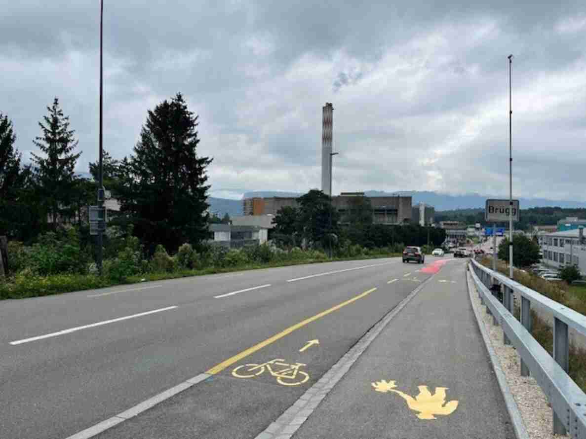 Strasse bei der Ortseinfahrt Brügg mit grüner Ortstafel am rechten Fahrbahnrand. Rechts führt ein baulich getrennter Radweg mit gelber Velo-Markierung und Richtungspfeil neben einem Leitplankengeländer. Auf dem Radbelag ist zudem ein gelbes Frosch-Symbol aufgemalt. Fahrzeuge nähern sich auf der Gegenfahrbahn, im Hintergrund ein Fabrikkamin und Industriegebäude vor Hügeln.