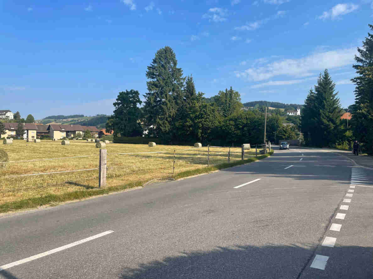 Kantonsstrasse in Blickrichtung Nord, angrenzend an ein Grundstück mit abgemähter Wiese und Heuballen, eingezäunt mit Elektrozaun. Die zweispurige Strasse mit Mittellinie und Fussgängerstreifen führt in einer Rechtskurve durch offenes Land Richtung Dorf mit Einfamilienhäusern und Kirchturm im Hintergrund. Ein einzelnes Auto fährt auf der Strasse, am rechten Strassenrand stehen zwei Personen.