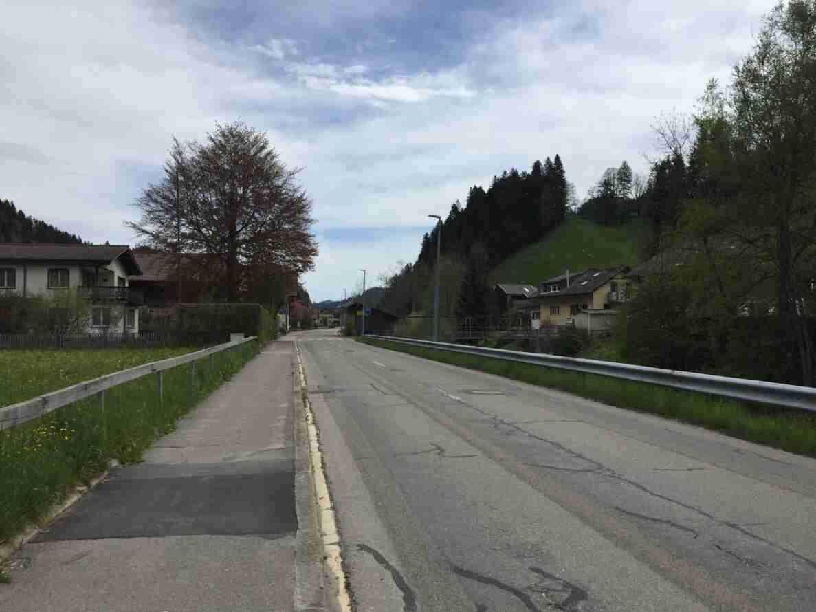Trubstrasse in Blickrichtung Nordosten, gerade verlaufende zweispurige Überlandstrasse mit angrenzendem Trottoir, das durch eine Holz- und Metallleitplanke von der Fahrbahn getrennt ist. Der Asphalt weist Risse und Flickstellen auf. Ein Fussgängerstreifen-Signal ist erkennbar. Die Strasse führt über eine kleine Brücke, rechts befindet sich eine Fussgängerbrücke über einen Bach. Rechts und links stehen Wohnhäuser, im Hintergrund bewaldete Hügel. Keine Personen oder Fahrzeuge sind auf der Strasse sichtbar.
