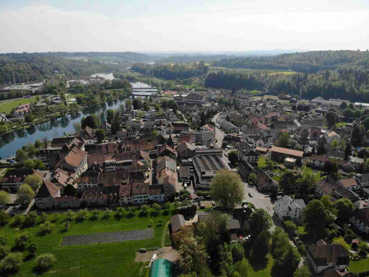 Drohnenaufnahme von Wangen an der Aare mit Blick auf den Fluss Aare, eine Brücke und die Kirche im Ortskern. Die Altstadt mit dichter Bebauung und roten Ziegeldächern erstreckt sich entlang mehrerer Strassen, die als Ortsdurchfahrten dienen. Der Strassenbelag ist trocken und intakt. Vereinzelt parkierte und fahrende Fahrzeuge sind auf den Strassen erkennbar, Fussgänger sind kaum sichtbar. Im Hintergrund Wald- und Landwirtschaftsflächen.