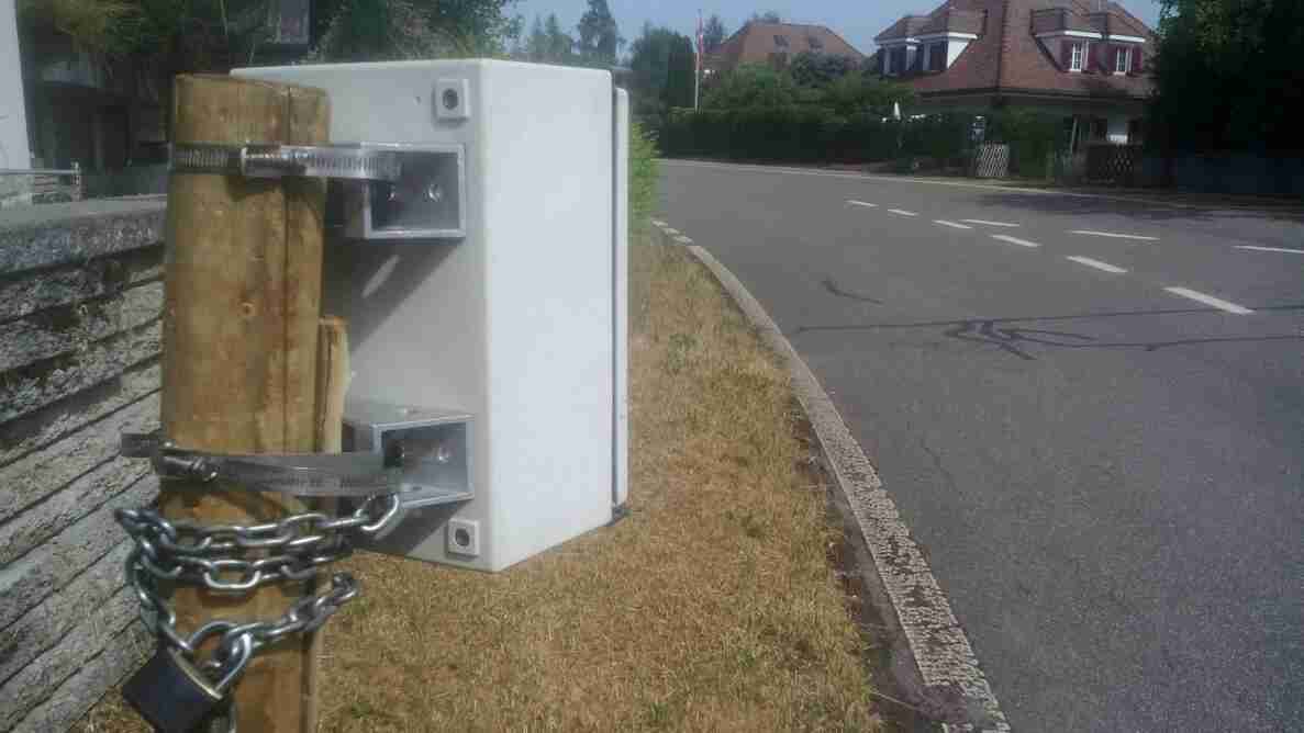 Weisses Messgerät, mit Schellen und Kette an einem Holzpfosten befestigt, am Rand einer geraden, asphaltierten Innerortsstrasse mit Mittellinie und Randmarkierung. Die Fahrbahn ist trocken und in gutem Zustand. Im Hintergrund eine leichte Rechtskurve, angrenzende Wohnhäuser mit Ziegeldächern, eine Schweizer Fahne sowie Bäume und Hecken säumen die Strasse.