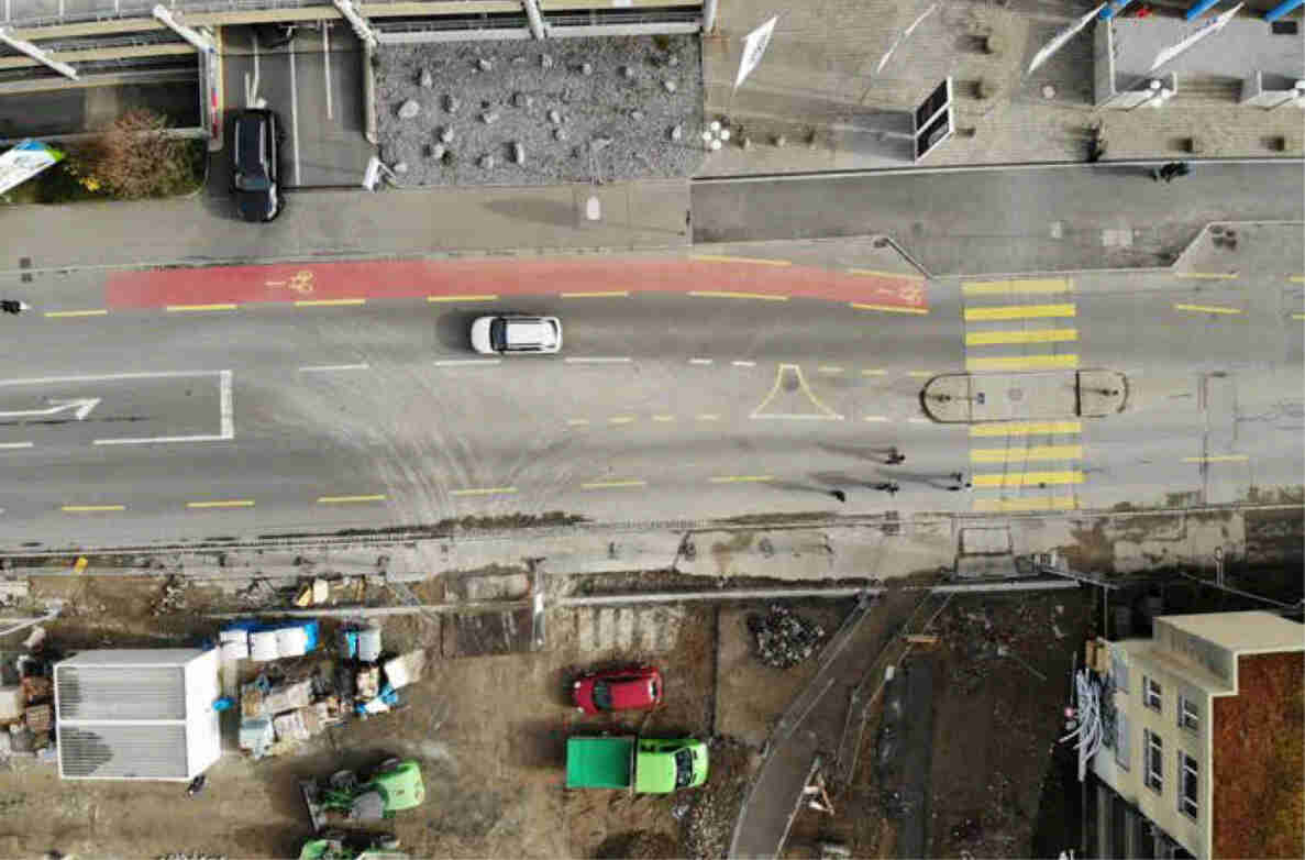 Vogelperspektive auf die Baslerstrasse mit Parkhauseinfahrt: zweispurige Fahrbahn mit rot markiertem Radstreifen, Mittelinsel und gelb-weisser Fussgängerstreifen-Markierung. Ein weisses und ein dunkles Auto befahren die Strasse, ein rotes Fahrzeug steht auf einem Nebenareal. Zwei Velofahrende überqueren die Fahrbahn. Der Belag ist teils uneben, angrenzend Baustellenmaterial und Container.