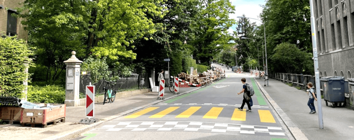 Gelb-weiss markierter Fussgängerstreifen mit schwarz-weissem Schachbrettmuster vor dem Schulhaus Münchhalde, auf dem ein Kind mit Rucksack die Strasse überquert. Rechts geht ein weiteres Kind auf dem Trottoir. Rot-weisse Leitbaken und Baustellenmaterial säumen den Fahrbahnrand, ein Fahrrad steht am Zaun angelehnt. Die Strasse ist asphaltiert und führt als ruhige Quartierstrasse zwischen Bäumen hindurch bergauf.