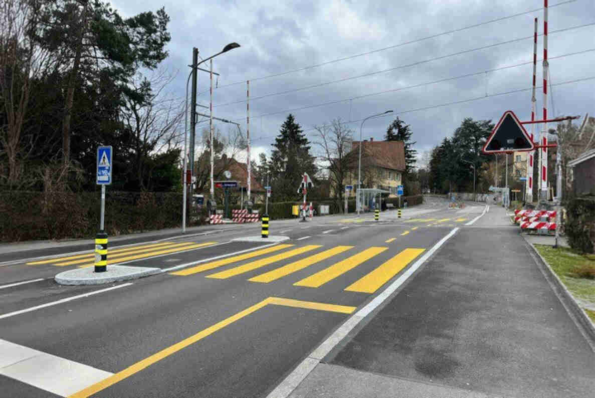 Bahnübergang Schweighof an der Schweighofstrasse in Zürich mit geschlossenen rot-weissen Barrieren beidseits der Gleise. Neu markierte gelbe Fussgängerstreifen mit Mittelinseln und Blinklichtsignalen queren die frisch asphaltierte Strasse. Ein Gefahrensignal für Bahnübergang sowie ein Hinweisschild für den Fussgängerstreifen stehen am Strassenrand. Die Fahrbahn ist beidseitig für den Verkehr freigegeben, aktuell sind keine Fahrzeuge oder Personen sichtbar.